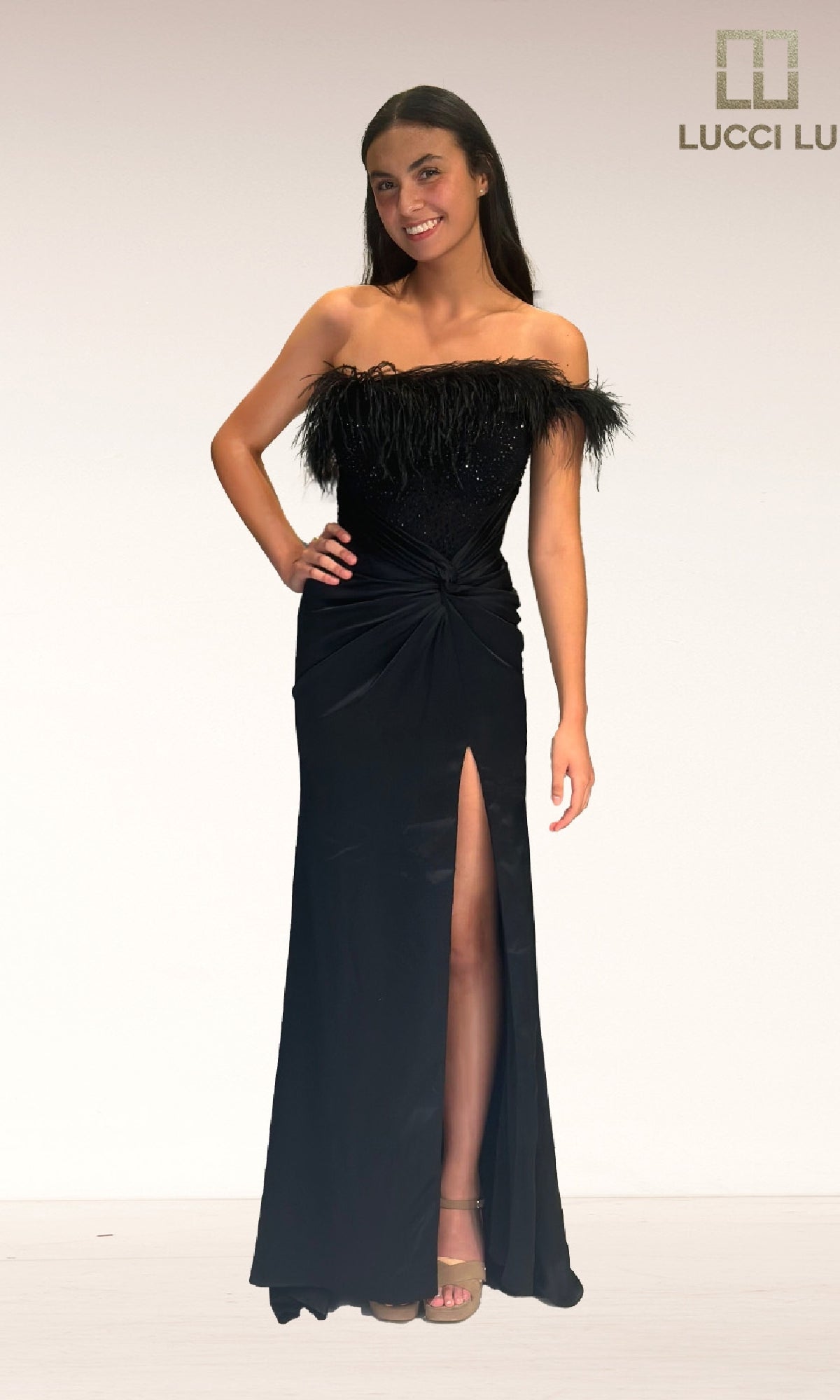 A woman in the Lucci Lu 1326 long prom dress, featuring black fabric, feather embellishments, an asymmetrical neckline, and a thigh-high slit, smiles against a plain background. The